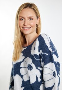 Blauw-witte gebreide trui met bloemenpatroon, zachte textuur, ronde halslijn en lange mouwen. Model draagt gouden hoepeloorbellen.
