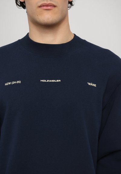 Námořnická modrá mikina s kulatým výstřihem, vyrobená z měkkého materiálu, s bílým textem: "HZW (24-25)," "HOLZWEILER," a "*MÅNE."