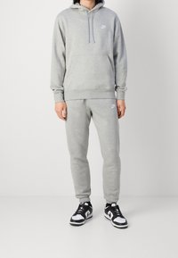 Person som bär en ljusgrå Nike-hoodie och sweatpants, i kombination med svarta och vita Nike-sneakers, står mot en enkel vit bakgrund.