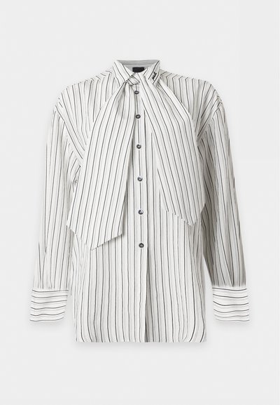 Blouse blanche à manches longues boutonnée avec fines rayures verticales noires et un grand détail de cravate asymétrique sur fond uni.