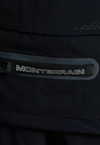 Κοντινή λήψη μαύρου φερμουάρ με υφασμάτινο τράβηγμα με την ετικέτα "MONTERRAIN" σε σκούρο ένδυμα.