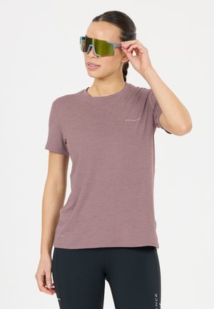 Vrouw die grote reflecterende sportzonnebril aanpast, draagt een mauve t-shirt en zwarte legging tegen een effen achtergrond.