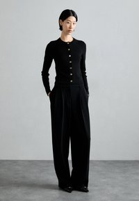 Svart cardigan med gullknapper, kombinert med skreddersydde svarte vide bukser. Glatt tekstur, tettsittende design og spisse svarte sko.