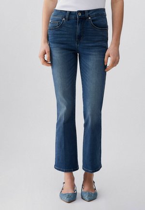 Persona che indossa jeans a vita media, blu scuro, a zampa e cropped con tasche frontali e chiusura con bottoni, abbinati a ballerine blu ricamate con punta appuntita.