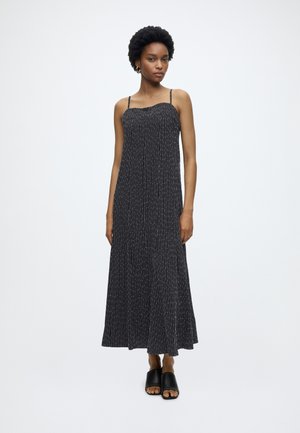 Femme aux cheveux courts et bouclés portant une robe maxi noire sans manches avec des rayures blanches verticales et des sandales slides noires à bout ouvert.