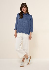 Blå skjorta med knappar och trekvartsärmar, scallopad nederkant och ögla-detaljer, i kombination med vita vida byxor och ljusa sneakers.