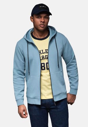 Sudadera con cremallera de color azul claro, con cordones, bolsillos frontales y puños de canalé, llevada sobre una camiseta amarilla con texto en azul marino.