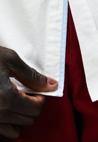 Biała koszula z niebieskim haftem, trzymana w dłoni. Koszula jest zestawiona z czerwonymi spodniami, z naciskiem na detale materiału i fakturę.