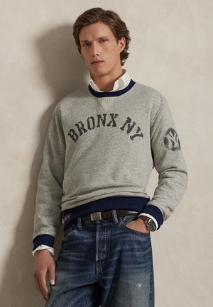 Gray sweatshirt featuring "BRONX NY" text and a logo on the sleeve. Navy accents on the collar and cuffs, paired with blue jeans.