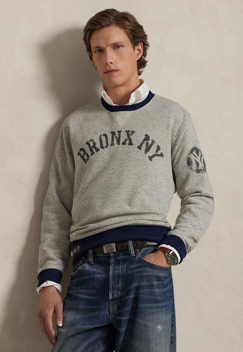 Gray sweatshirt featuring "BRONX NY" text and a logo on the sleeve. Navy accents on the collar and cuffs, paired with blue jeans.