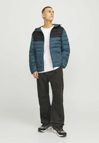 Chaqueta acolchada en verde azulado y negro, con cierre frontal, capucha y costuras horizontales. Lleva encima una camiseta blanca y combinada con pantalones negros.