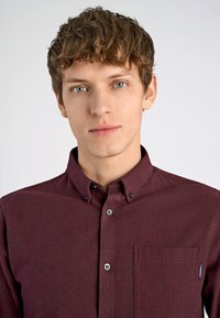 Bordeaux knoopshirt van een gestructureerde stof, korte kraag, zak aan de linkerkant en donkere knopen. Bevat zichtbare stikseldetails.