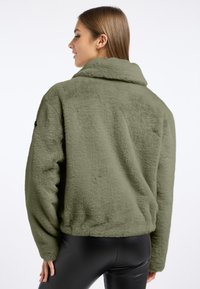 Futrzana oliwkowa kurtka z kołnierzem i elastycznym dołem; posiada długie rękawy oraz teksturowany materiał. Akcenty czarnych elementów metalowych na rękawie.