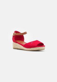 Punainen viistokärkinen sandaali, jossa on avoin kärki, tanvärinennilkkaremmi soljella ja punottu espadrille-tyylinen pohja valkoista taustaa vasten.
