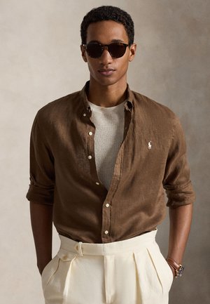 Άνδρας με σκούρα γυαλιά ηλίου, καφέ πουκάμισο με κουμπιά πάνω από λευκό υφασμάτινο πουλόβερ, εκρού πλισέ παντελόνια και ρολόι χειρός, σε ουδέτερο φόντο.