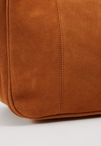 Gros plan d'un coin de sac en suède marron montrant une texture douce et des coutures détaillées sur un tissu lisse.