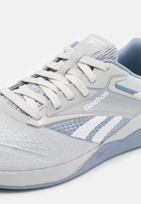 Light gray athletic shoe with mesh upper, tonal laces, and blue accents. Features Reebok logo and textured sole with contrasting stripes.