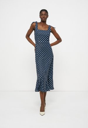 Kobieta ubrana w granatową sukienkę midi w białe groszki, związaną na ramiączkach, z falbaną u dołu, pozuje z rękami na biodrach, w beżowych szpilkach.
