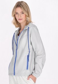 Giacca a vento grigia chiara con zip e cordini blu, dotata di cappuccio e polsini elastici, realizzata in un materiale liscio e leggero.
