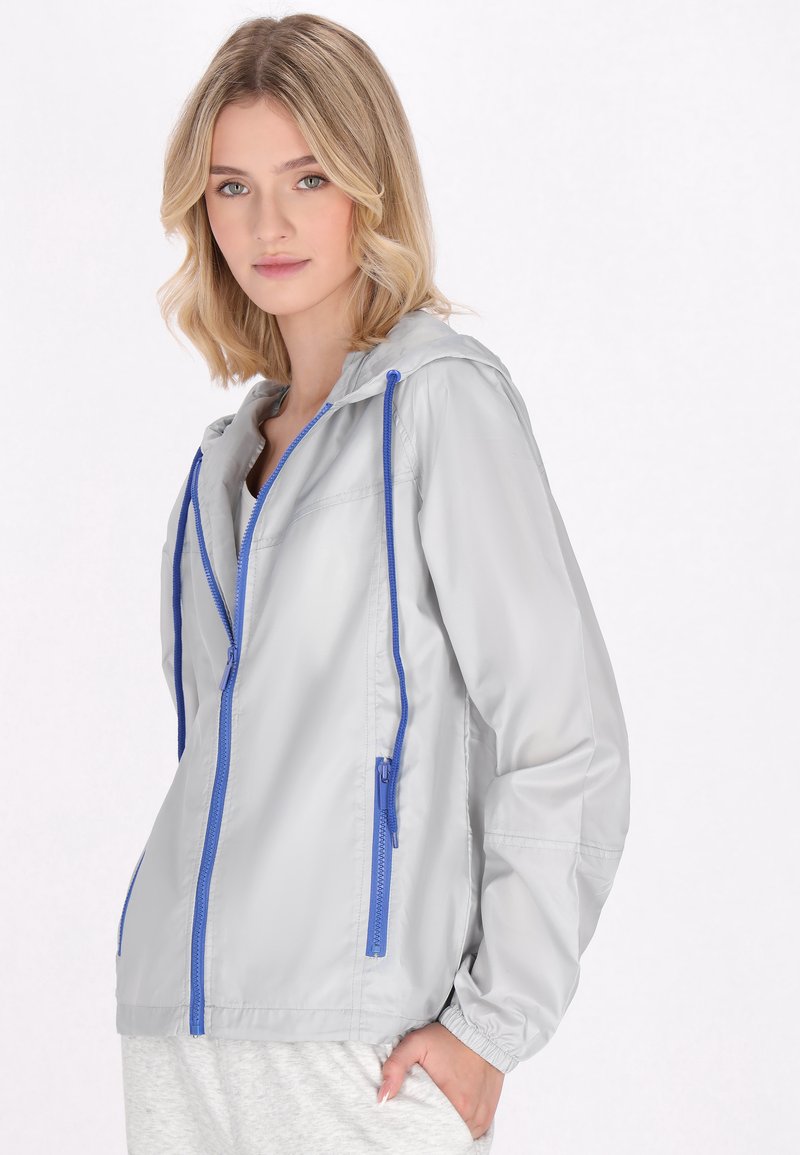 Giacca a vento grigia chiara con zip e cordini blu, dotata di cappuccio e polsini elastici, realizzata in un materiale liscio e leggero.