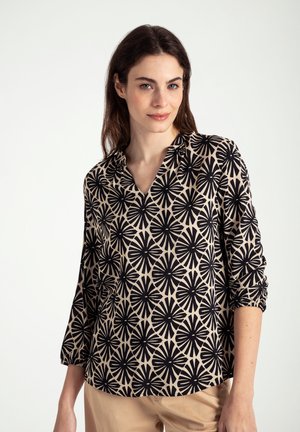 Zwarte blouse met bloemenpatroon op een lichtbeige achtergrond, met een v-halslijn en driekwartmouwen. Zachte stofstructuur.