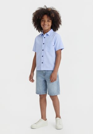 Αγόρι με σγουρά μαλλιά που χαμογελάει, φοράει μπλε ανοιχτό κοντομάνικο πουκάμισο με κουμπιά, τζιν σορτς και λευκά αθλητικά παπούτσια, στέκεται σε λευκό φόντο.