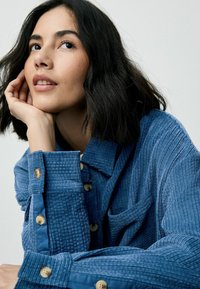 Camicia in corduroy blu con design a bottoni, presenta una tasca, tessuto strutturato e colletto arrotondato. I bottoni sono di colore marrone chiaro.