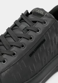 Sapatilha de couro preto com detalhes em borracha texturizada e atacadores correspondentes. Apresenta um logótipo gravado e detalhes de costura em contraste.