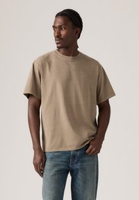 Mann med flettehår iført en beige kortermet Levi's t-skjorte og blå jeans, ser mot venstre mot en enkel bakgrunn.