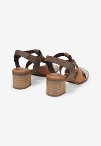 Sandalias de ante marrón con tacón de madera gruesa, que cuentan con detalles en las correas y un diseño abierto. Piso texturizado visible.