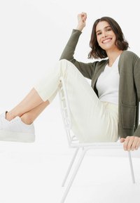 Olivgrüner Cardigan über einem weißen Hemd, kombiniert mit cremefarbenen Hosen und weißen Sneakers, sitzt auf einem weißen Stuhl vor einem schlichten Hintergrund.