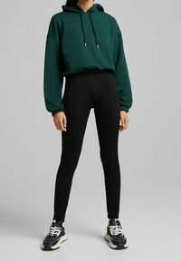 Sudadera verde con cordones, puños acanalados; leggings negros de talle alto; combinados con zapatillas blancas y negras con suela gruesa.