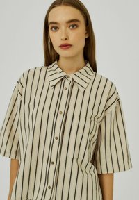 Gestreept, kortärmig shirt in beige en zwart. Heeft een klassieke kraag, knoopsluiting en een borstzak. Zachte stoftextuur.