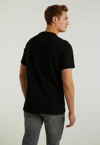 Tricou negru din bumbac, cu mâneci scurte, guler rotund și o croială lejeră, văzut din spate. Materialul pare neted, cu o întindere minimă.