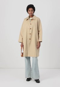 Trench beige con colletto e bottoni frontali. Dotato di tasche laterali, vestibilità rilassata ed è abbinato a jeans chiari a zampa d'elefante.