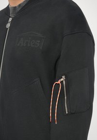 Sudadera negra con cierre de cremallera, que presenta un logo texturizado de "Aries", un bolsillo con cremallera en la manga y un cordón naranja y blanco.
