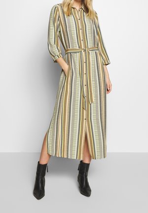 Nainen, jolla on päällään pitkä raidallinen nappimekko vyöllä, kolmanneksen pituisilla hihoilla, sivuhalkioilla ja mustat nilkkurit, yksinkertaisen taustan edessä.