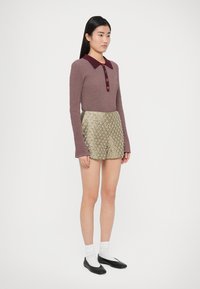 Langärmeliger Rippsweater in Mauve mit einem kastanienbraunen Kragen und Knöpfen, kombiniert mit metallisch goldfarbenen Shorts mit diamantförmigem Muster.