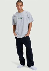 T-shirt cinza oversized de algodão com mangas curtas, apresentando um logo gráfico verde, combinado com calças largas azul-marinho e ténis brancos.