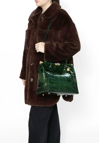 Borsa a tracolla in pelle verde con motivo coccodrillo, dotata di hardware dorato, forma strutturata e tracolla removibile, portata (tenuta) con un cappotto marrone.