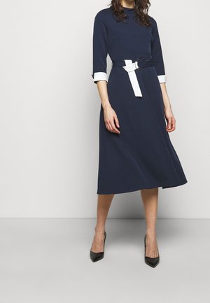 Femme portant une robe midi bleu marine avec des poignets blancs et une ceinture blanche, associée à des escarpins noirs à bout pointu, debout contre un mur blanc.