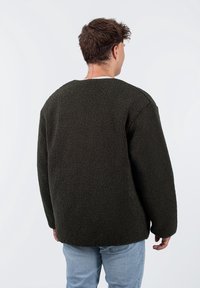 Ciemnozielona polarowa kurtka o fakturowanej powierzchni, okrągłym dekolcie, luźnym kroju i opadających ramionach, noszona na jasnych dżinsach.