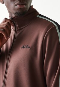 Καφέ τζάκετ με φερμουάρ και ψηλό γιακά, με μαύρες και ανοιχτοπράσινες ρίγες ως διακοσμητικά στοιχεία. Κεντημένο λογότυπο "New Era" στο στήθος.