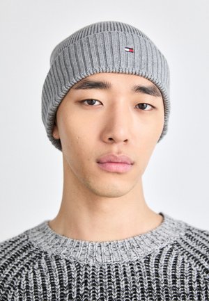 Gray ribbed knit beanie with a small logo in red and blue. Worn by a person in a contrasting gray sweater with a textured pattern.