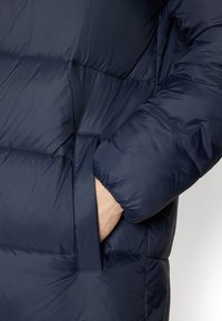 Primer plano de una chaqueta de plumón azul marino con diseño acolchado, textura suave y detalle de bolsillo lateral con puños elásticos en las mangas.