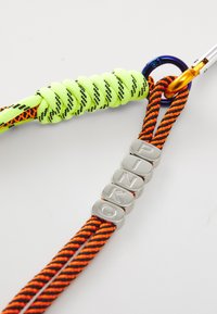 Šarena vezica ima pleteni neon zelenu žicu i narančaste pruge, sa srebrnim slovima koja oblikuju riječ "PINKO" i metalni karabiner.