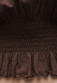 Σκούρο καφέ ύφασμα με συγκεντρωμένο ελαστικό σούρωμα και λεπτομέρεια με βολάν στο τελείωμα.