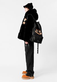 Abrigo de piel negra con bordado de oso de peluche, mochila negra, pantalones gris claro con detalles de herrajes y zapatos plataforma marrones.