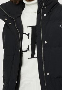 Sort puffer vest med høj krave, trykknapper og lynlås. Har et lille logo patch på brystet, båret over en hvidmønstret sweater.