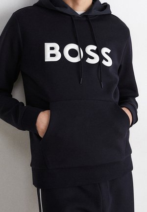 Osoba nosząca czarną bluzę z kapturem z białymi sznureczkami i dużymi białymi literami tworzącymi napis "BOSS" na klatce piersiowej, ręce w przedniej kieszeni.